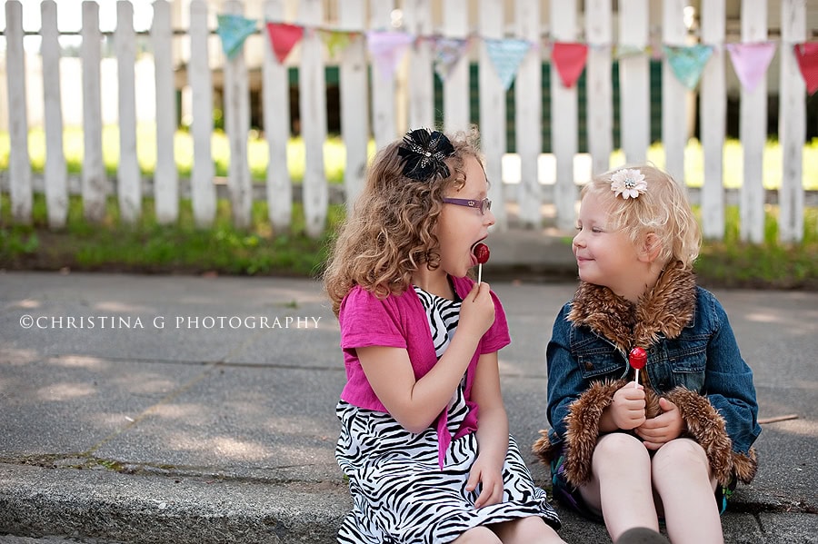 TWO LITTLE GIRLS SITTING ON SIDEWALK WITH LOLLIPOPS
